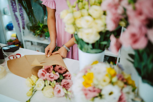 Κοντινό πλάνο των χεριών μιας καυκάσιας γυναίκας με ένα ροζ φόρεμα που οργανώνει ένα μπουκέτο λουλούδια σε ένα τραπέζι σε ένα ανθοπωλείο. Φτιάχνω ένα μπουκέτο λουλούδια - Φωτογραφία, εικόνα