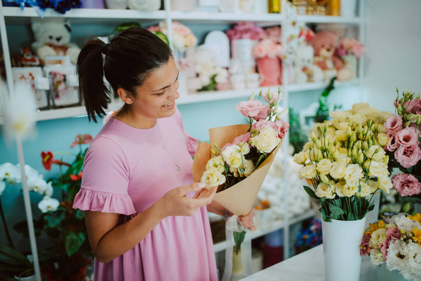 Jeune jolie brune caucasienne fleuriste dans une robe rose arrangeant un bouquet de fleurs dans un magasin de fleurs - Photo, image