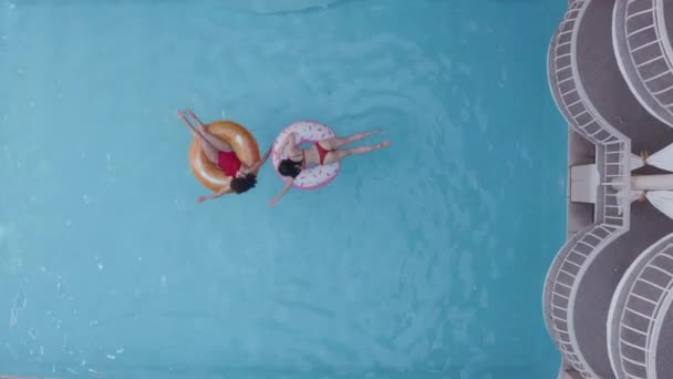 Daleko natáčení shora ze dvou brunetek plavání v modrém bazénu na nafukovací kruhy dotknout vody mluví relaxační. Dvě sestry spolu tráví čas v gumových kroužcích a opalování v hotelovém komplexu - Záběry, video
