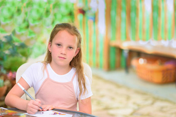 Little freckles girl painting at table in summer garden. Serious kid drawing outdoor and looking at camera. Cute child doing homework at sunny yard.Copy space for text. - Fotó, kép