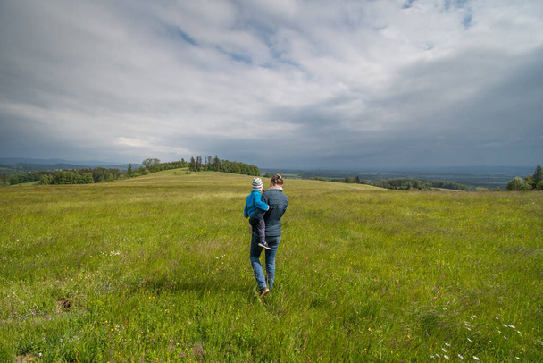 Anne kucağında bebekle vadiye ve bulutlu gökyüzüne bakan yeşil bir çayırda yürüyor. - Fotoğraf, Görsel