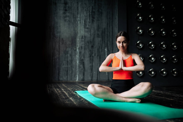 Portrait of a meditating yoga trainer girl in lotus position who folded her hands in a prayer gesture in front of her chest sitting on a wooden floor - Foto, Imagem