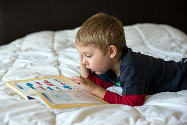 een jonge blonde jongen is het leren op een bed - Foto, afbeelding
