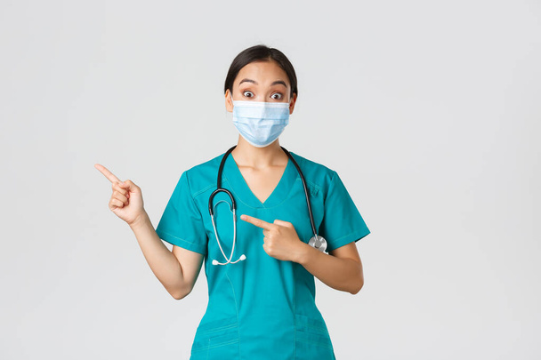 Covid-19, coronavirus disease, healthcare workers concept. Surprised and interested asian female doctor, physician in medical mask and scrubs pointing fingers upper left corner, white background - Photo, Image