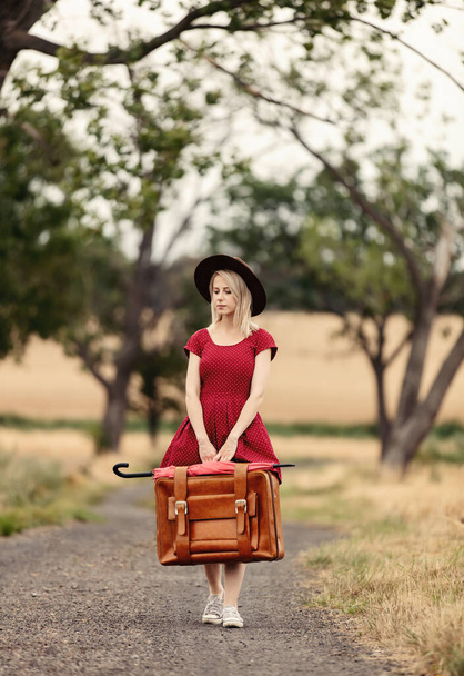 blonde in red dress with a suitcase on a rural road before the rain - Foto, Bild