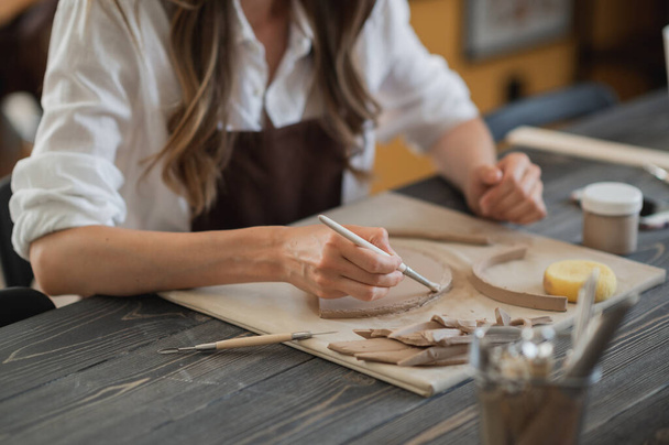 As mãos do artesão conectam as peças de argila feitas de argila crua, os dedos principais trabalham com pedaços de argila, a fêmea senta-se em uma oficina atrás da mesa - Foto, Imagem