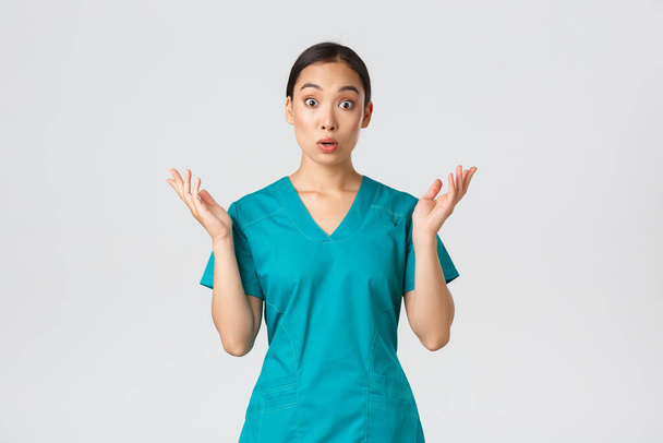 Covid-19, healthcare workers, pandemic concept. Surprised and amazed asian nurse in scrubs raising hands up, looking startled, found out big news, hospital got coronavirus vaccine, white background - Photo, Image
