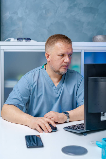 Doctor in blue scrubs sitting near computer. Searching medical information. Diagnostic and medical concept. - Foto, Imagem