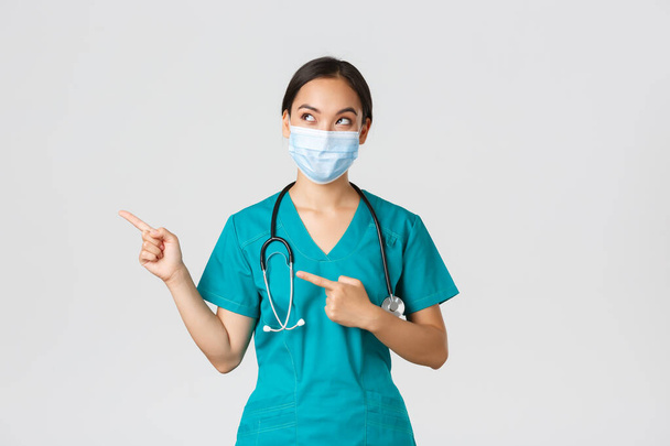 Covid-19, coronavirus disease, healthcare workers concept. Curious pleased asian female doctor, physician in medical mask and gloves, looking and pointing upper left corner with intrigued smile - Photo, Image