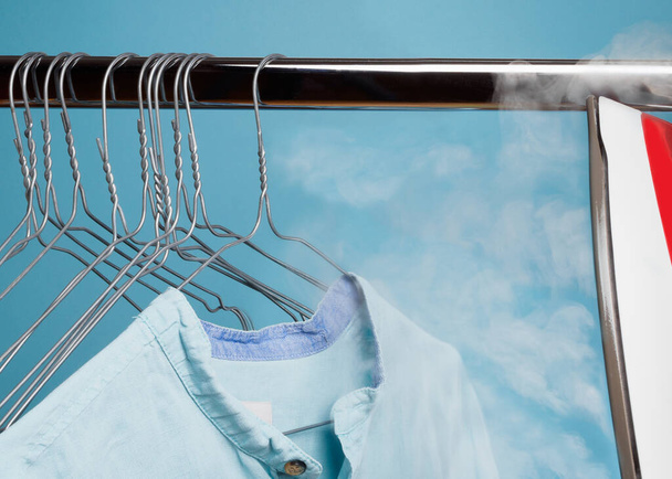 a blue shirt hangs on the shoulders and is steamed by the steam of an iron on a blue background - Photo, Image