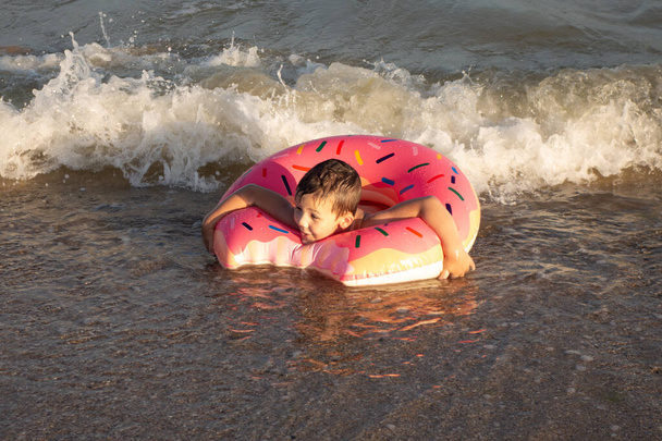 Веселый мальчик 5 лет плавает в море на надувном кольце в виде пончика в солнечный день. - Фото, изображение