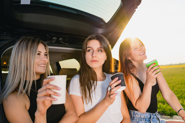 Three girls have coffee break at car trunk. Happy road trip smiling female travelers, relax at vehicle back with coffee cups take away at sunlight. - Photo, Image