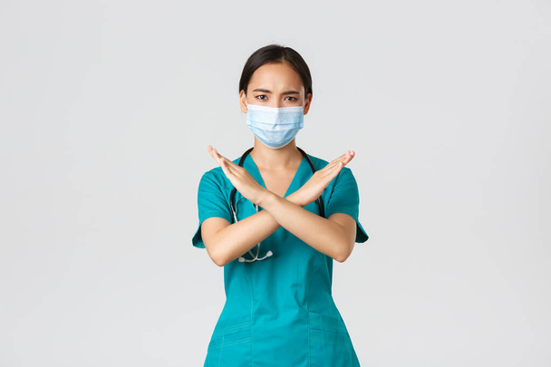 Covid-19, coronavirus disease, healthcare workers concept. Serious displeased, frowning asian doctor in scrubs show cross gesture in disapproval, stop someone, prohibit action, white background - Photo, Image