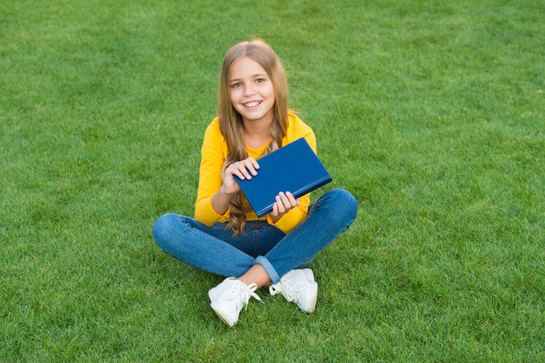 Girl reading book outdoors green lawn background, kids literature concept - Foto, imagen