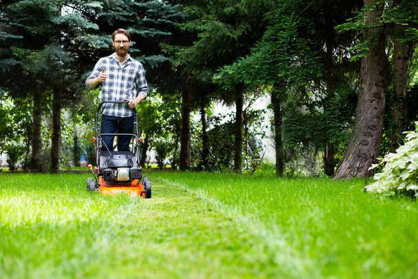 Gardening. Greenworks. Man with beard mows grass using a petrol lawnmower with adjustable cutting heights. Mowing a lush, leafy green lawn by motor mower, gasoline lawnmower in a garden on a sunny day - Foto, Bild