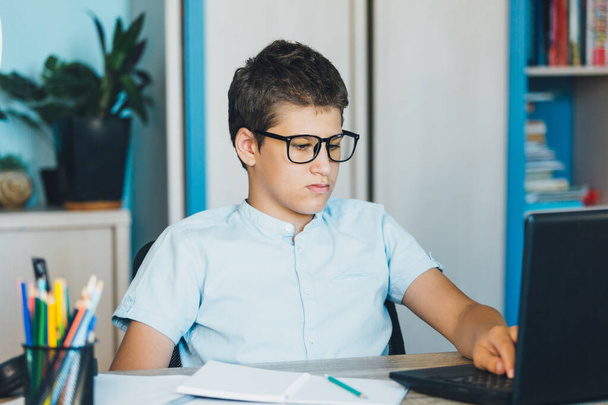 Cute young boy in blue shirt sitting behind desk in his room next to laptop and study. Teenager in earphones makes homework, listening lesson. Home, distance education, self education by  kids. - 写真・画像