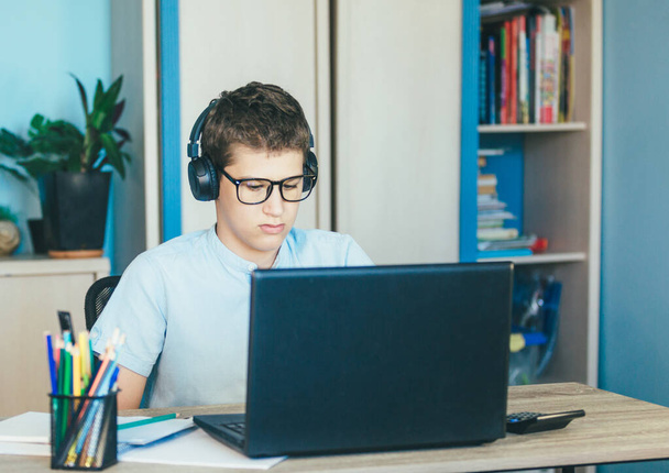 Uroczy, młody chłopak w niebieskiej koszuli siedzi za biurkiem w pokoju obok laptopa i uczy się. Nastolatek w słuchawkach odrabiał lekcje, słuchał. Dom, edukacja na odległość, samokształcenie przez dzieci. - Zdjęcie, obraz