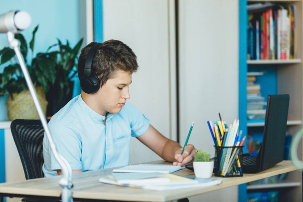 Милий молодий хлопчик у блакитній сорочці сидить за столом у своїй кімнаті поруч з ноутбуком та навчанням. Підліток в навушниках робить домашнє завдання, прослуховуючи урок. Будинок, дистанційна освіта, самоосвіта дітей
. - Фото, зображення