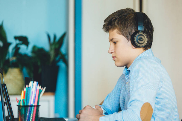 Aranyos, kék inges fiú ül az íróasztal mögött a laptopja mellett és tanul. A fülhallgatós tinédzser házi feladatot csinál, hallgatózik. Otthon, távoktatás, gyermeknevelés. - Fotó, kép