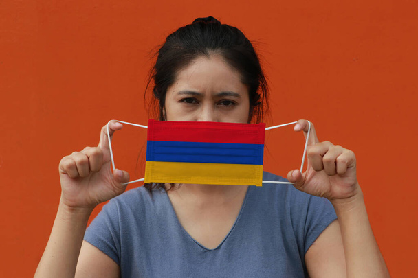 Kobieta z ormiańską flagą na masce higienicznej w ręku podniosła twarz na pomarańczowym tle. Drobne cząstki lub korona wirusa lub ochrona Covid 19. Koncepcja zwalczania chorób. - Zdjęcie, obraz
