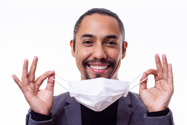 close-up van een latijn man glimlachen gelukkig terwijl het houden van een medisch masker met beide handen op witte achtergrond. - Foto, afbeelding