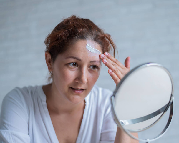 Portrait of a Caucasian woman with vitiligo uses sunscreen. A girl with a white pigment spot on her forehead looks in the mirror and is smeared with cream. - Zdjęcie, obraz