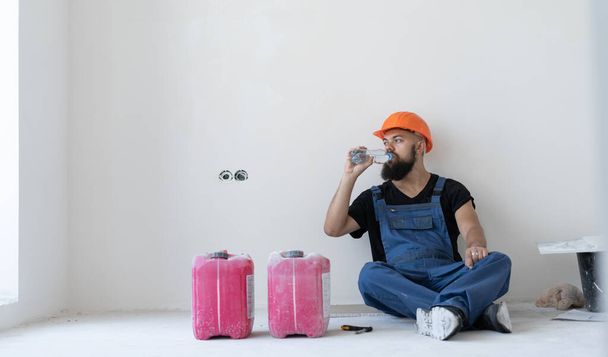Ruhe beim Erbauer. Ein bärtiger Mann in blauer Uniform und orangefarbenem Hut sitzt während einer Pause auf dem Boden. Wasser aus der Flasche trinken. Platz für Text. Kopierraum. - Foto, Bild