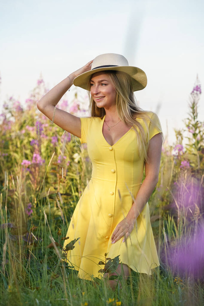 Outdoors portrait of beauty blond woman in summer clothes. Young model in hat and yellow dress posing at flowers field at daytime  - Fotografie, Obrázek
