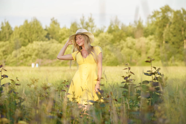 Outdoors portrait of beauty blond woman in summer clothes. Young model in hat and yellow dress posing at flowers field at daytime  - Foto, Imagen