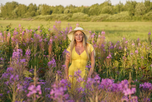 Outdoors portrait of beauty blond woman in summer clothes. Young model in hat and yellow dress posing at flowers field at daytime  - Zdjęcie, obraz