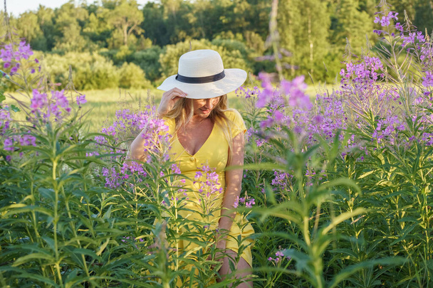 Outdoors portrait of beauty blond woman in summer clothes. Young model in hat and yellow dress posing at flowers field at daytime  - Foto, immagini
