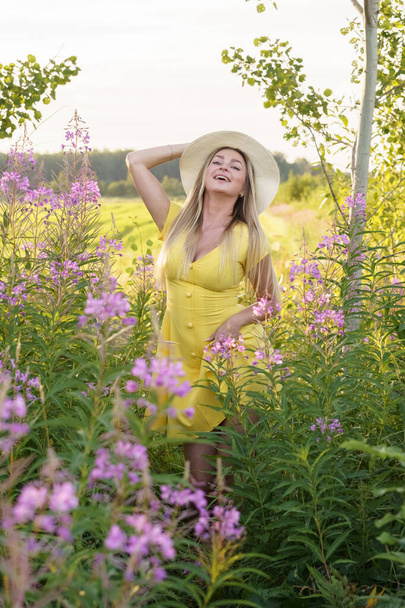 夏服の美しさブロンドの女性の屋外肖像画。昼間の花畑でポーズをとる帽子と黄色のドレスの若いモデル  - 写真・画像