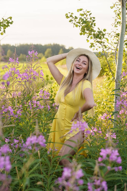 Outdoors portrait of beauty blond woman in summer clothes. Young model in hat and yellow dress posing at flowers field at daytime  - Photo, image
