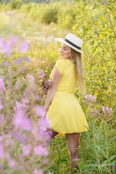 Outdoors portrait of beauty blond woman in summer clothes. Young model in hat and yellow dress posing at flowers field at daytime  - Фото, изображение