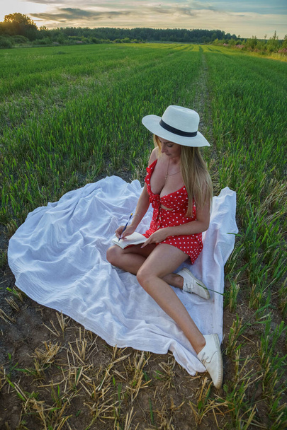 Outdoors portrait of beauty blond woman in summer clothes. Young model in hat and red dress resting at flowers field at daytime  - Fotoğraf, Görsel