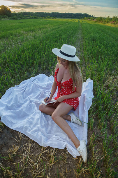 Outdoors portrait of beauty blond woman in summer clothes. Young model in hat and red dress resting at flowers field at daytime  - Zdjęcie, obraz