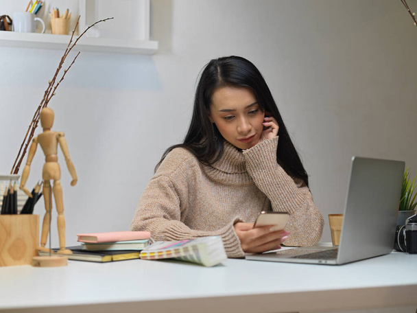 Portrait of female office worker sitting at office desk while using smartphone in office room - 写真・画像
