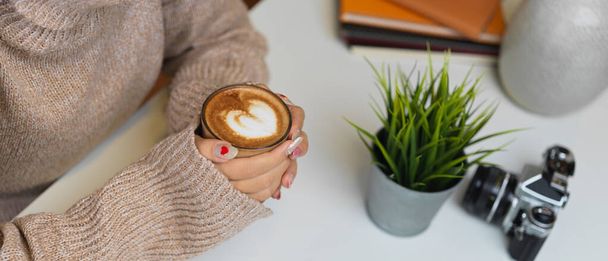 Side view of female in sweater hands holding a cup of coffee on table with camera and supplies - Photo, Image