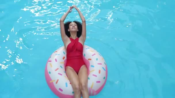 Etnické kudrnaté ženy vznášející se v modré vodě v hotelovém bazénu v nafukovací trubici zvednou ruce nad hlavu a usmějí se na kameru. Afro student dívka relaxovat v turistickém komplexu plavání na kruhu - Záběry, video