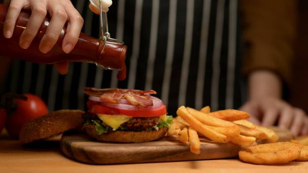 Vista de cerca de la mano femenina con una botella de ketchup añadiendo en fresco sabroso tocino hamburguesa en la cocina  - Foto, imagen