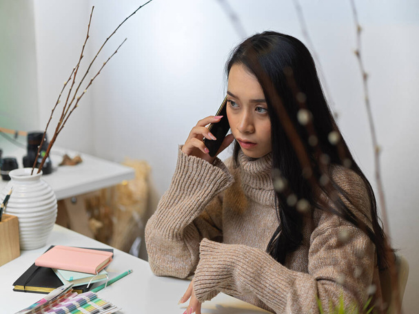 Καλλιεργημένη φωτογραφία της γυναίκας σε πουλόβερ μιλάμε στο τηλέφωνο, ενώ κάθεται στο τραπέζι εργασίας με γραφική ύλη, προμήθειες και διακοσμήσεις - Φωτογραφία, εικόνα