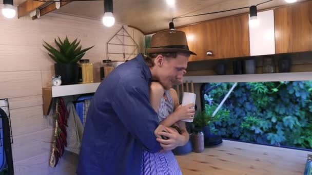 Positive young couple living or travelling in van. Smiling, stylish couple standing embracing in kitchen, woman gives her husband the tea box to smell it. Stylish, cozy interior - Séquence, vidéo