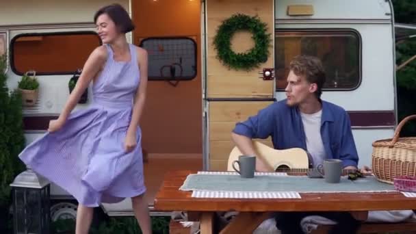 Funny girl in blue summer dress girl dancing while man playing guitar sitting at the wooden table in front trailer, singing a song loudly. Vacation, holidays, trailer trip - Footage, Video