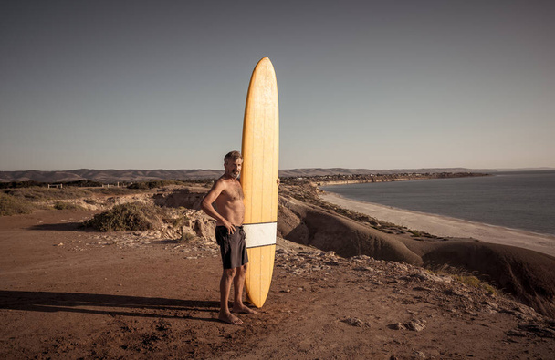 Attractive Australian mature man surfer with cool vintage surfboard on beach at sunset. Senior adult happy to be Back to surf . Outdoors sports adventure, active older people and retirement lifestyle. - Foto, Bild