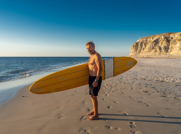 Attractive Australian mature man surfer with cool vintage surfboard on beach at sunset. Senior adult happy to be Back to surf . Outdoors sports adventure, active older people and retirement lifestyle. - Photo, Image