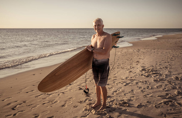 Attractive Australian mature man surfer with cool vintage surfboard on beach at sunset. Senior adult happy to be Back to surf . Outdoors sports adventure, active older people and retirement lifestyle. - Valokuva, kuva