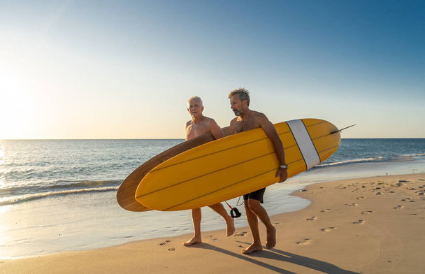Two mature men walking with surfboards on beautiful beach enjoying paradise and retirement lifestyle. Attractive fit senior adults friends having fun surfing. In real active and healthy people. - Fotografie, Obrázek