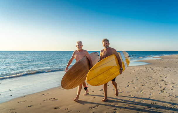 Two mature men walking with surfboards on beautiful beach enjoying paradise and retirement lifestyle. Attractive fit senior adults friends having fun surfing. In real active and healthy people. - Fotografie, Obrázek