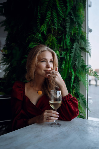 vrouw met glas witte wijn in café of restaurant. sexy jong blond meisje. concept van feesten, genieten van alcohol, stijl en modieuze levensstijl. Portret meisje in rode jurk met wijnglas - Foto, afbeelding
