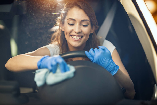 Μια νεαρή χαμογελαστή γυναίκα με προστατευτικά γάντια στα χέρια της, σκουπίζει ένα τιμόνι με ένα πανί έτοιμο να χτυπήσει το δρόμο.  - Φωτογραφία, εικόνα